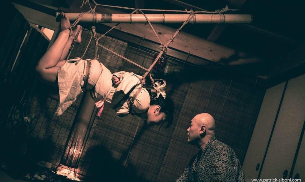 Dark room with low light, woman suspended in rope bondage from a bamboo stick with a man looking at his work of art.